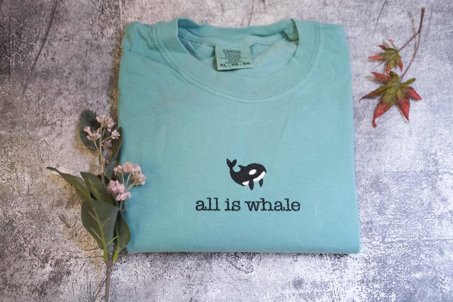 A light blue t-shirt with a black and white whale graphic and the text &quot;all is whale&quot; printed on it, along with some flowers and a fallen leaf on a textured gray surface.