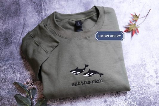 a green t-shirt with a black and white whale design and the text "eat the rich" printed on it, placed on a gray surface with some leaves and flowers.