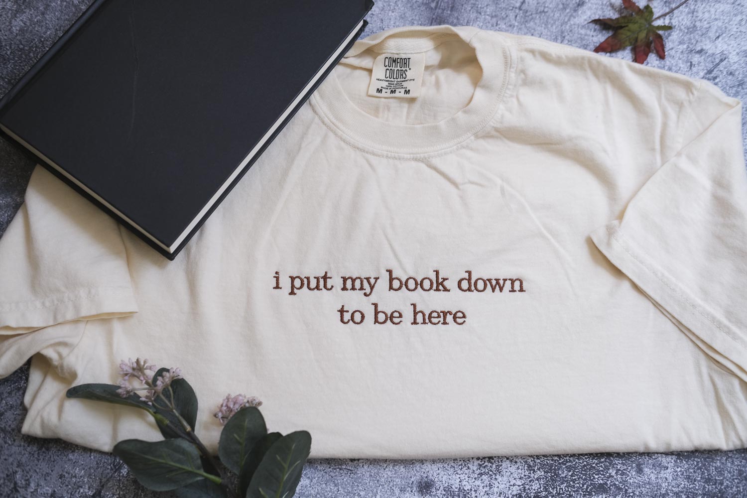 a white t-shirt with the text "I put my book down to be here" printed on it, along with a black book and some flowers.