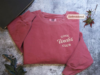 A red long-sleeved shirt with the text "Cool Uncle Club" embroidered on it, along with a leaf and a book, all placed on a gray surface.