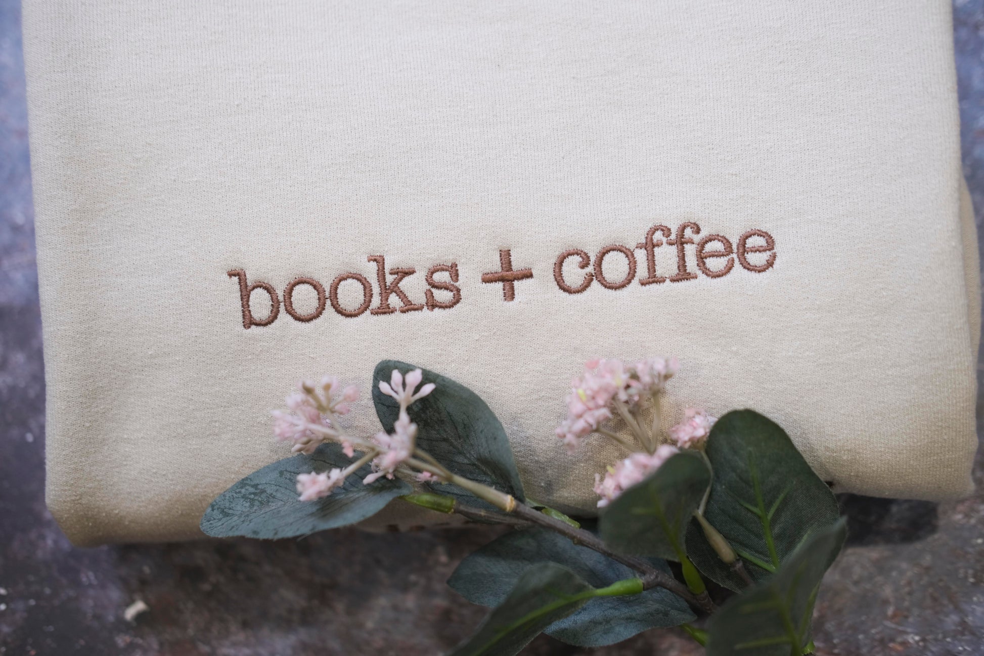 a beige fabric with the text "books + coffee" embroidered on it, along with a small branch of pink flowers and green leaves.