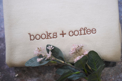 a beige fabric with the text "books + coffee" embroidered on it, along with a small branch of pink flowers and green leaves.