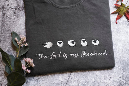 A black t-shirt with a white sheep design and the text "The Lord is my Shepherd" printed on it, along with some leaves and flowers.