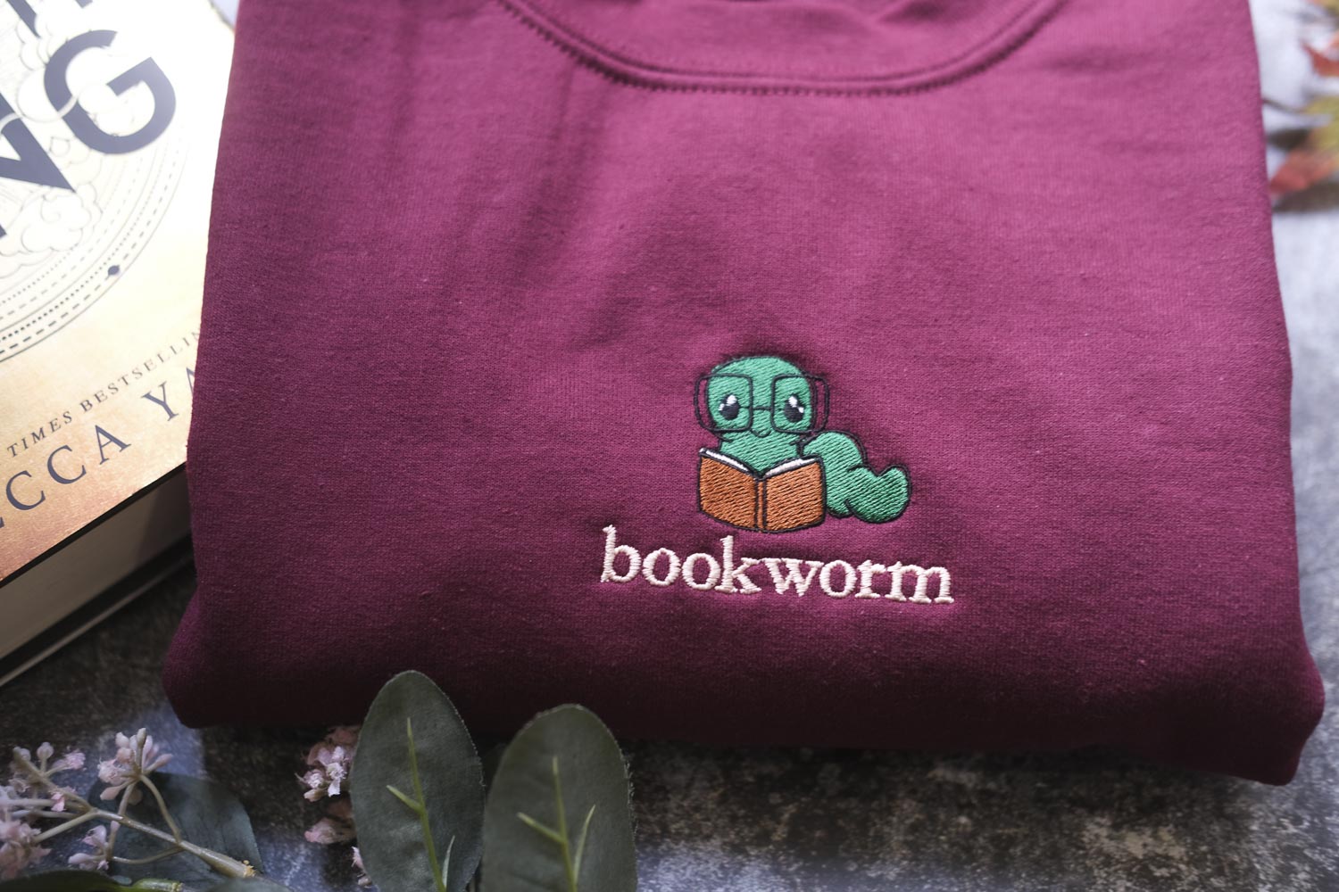 A maroon sweatshirt with a small green book worm embroidered on it, along with a book and some flowers.