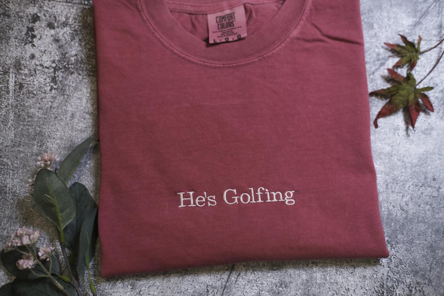 A red t-shirt with the text "He's Golfing" printed on it, along with some leaves and flowers.