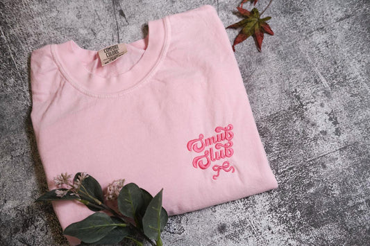 a pink t-shirt with a pink embroidered logo, placed on a gray surface alongside some leaves and a small red leaf.