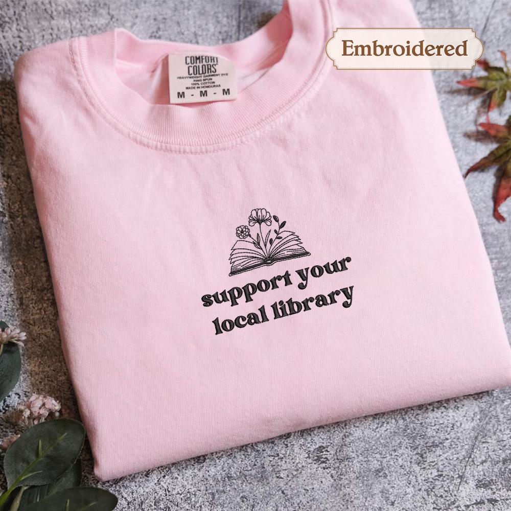 A pink t-shirt with the text &quot;Support your local library&quot; printed on it, along with an embroidered design of an open book and flowers.