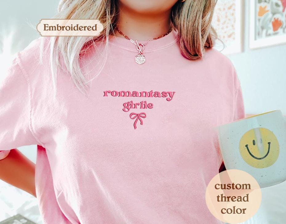 A person wearing a pink t-shirt with the text &quot;romancey girl&quot; embroidered on it, holding a mug with a smiley face on it.