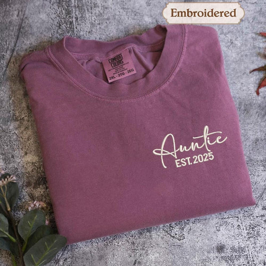 A purple t-shirt with the text &quot;Auntie EST.2025&quot; embroidered on it, along with a label indicating it is &quot;Embroidered&quot; and &quot;Comfy Colors&quot; in the neck area.