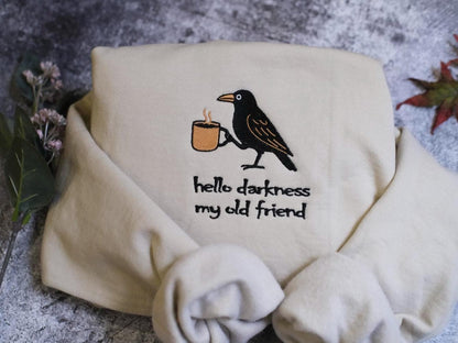 A white sweatshirt with a black bird embroidered on it, holding a cup of coffee, and the text &quot;Hello darkness, my old friend&quot; printed on it.