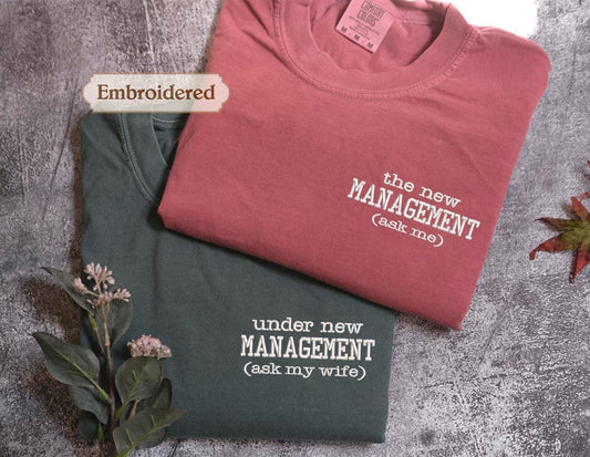 Two t-shirts are displayed, one in a dark green color and the other in a red color, both with humorous text printed on them.