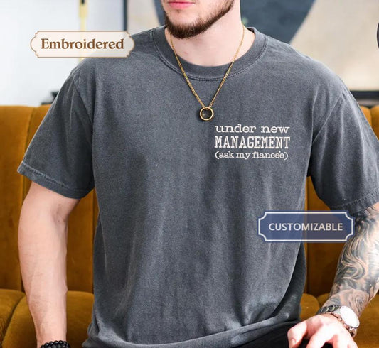 A man wearing a gray t-shirt with the text &quot;under new MANAGEMENT (ask my fiancée)&quot; embroidered on it.