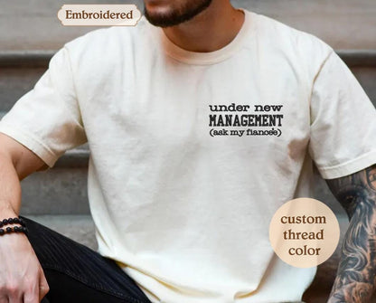 A man wearing a white t-shirt with the text &quot;under new MANAGEMENT (ask my flirts)&quot; printed on it.
