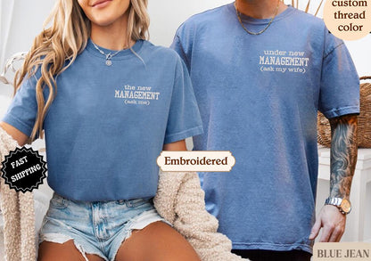 A woman wearing a blue t-shirt with the text &quot;the new MANAGEMENT (ask me)&quot; and a man wearing a blue t-shirt with the text &quot;under new MANAGEMENT (ask my wife)&quot; standing next to each other.