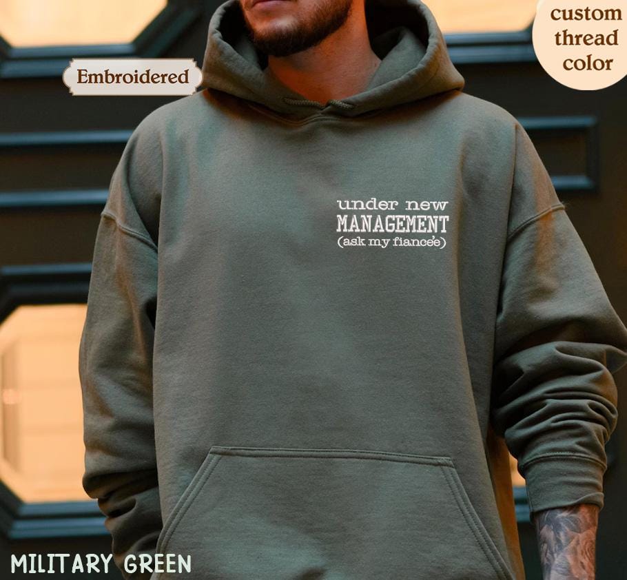 A person wearing a green hoodie with the text &quot;under new MANAGEMENT (ask my fiancée)&quot; printed on it.
