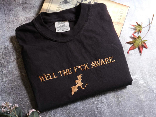 a black t-shirt with gold text and a graphic design, placed on a gray surface alongside some leaves and a book.