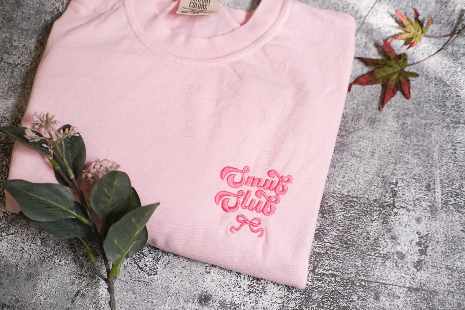 A pink t-shirt with a pink embroidered text on it, along with some leaves and a small flower, placed on a gray surface.