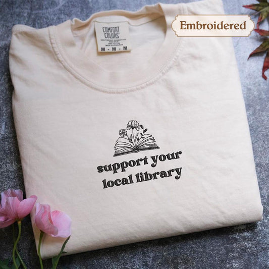 A white t-shirt with a printed message on it, placed on a gray surface with pink flowers nearby.