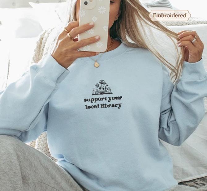 A woman is sitting on a bed, holding a phone up to her face, taking a selfie. She is wearing a light blue sweater with a message printed on it.
