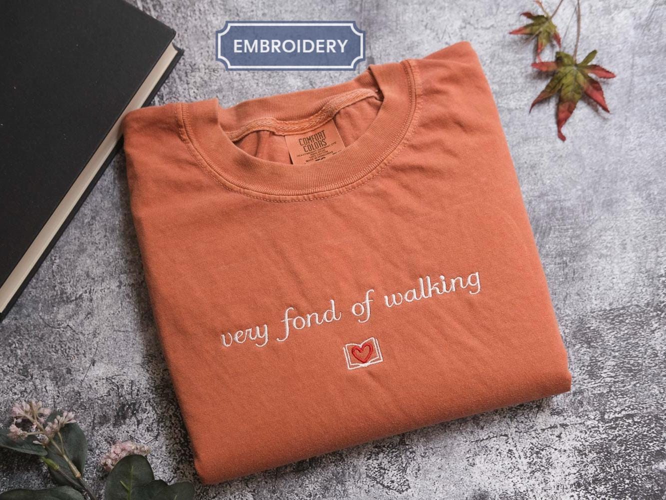 a brown t-shirt with the text &quot;very fond of walking&quot; printed on it, along with a small red heart symbol. The t-shirt is folded and placed on a gray surface, with a black book and some leaves in the background.