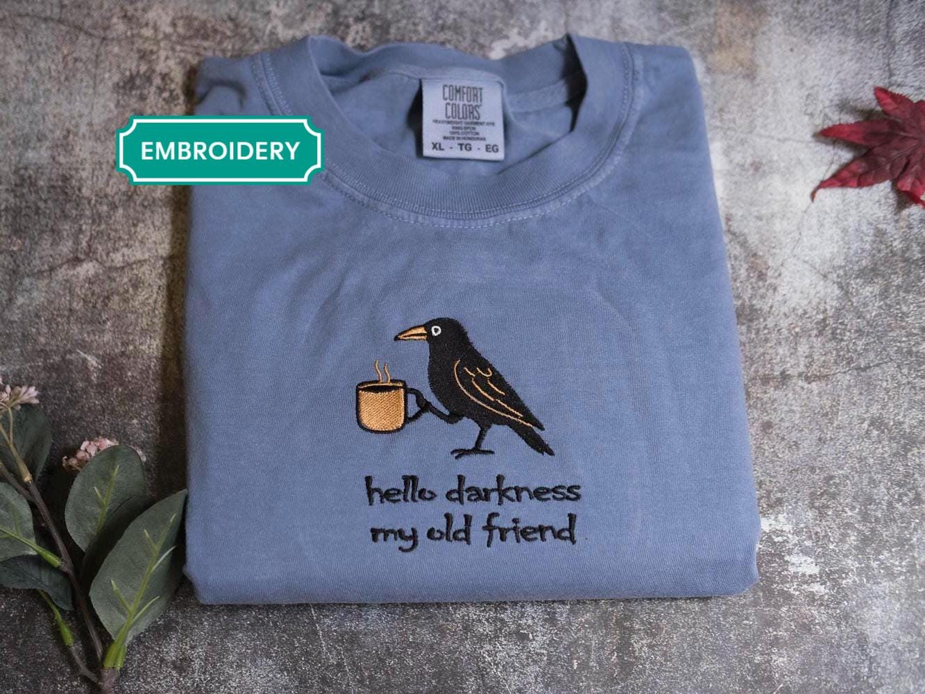 A gray t-shirt with a black bird holding a cup of coffee, accompanied by the text &quot;Hello darkness, my old friend&quot; printed on it.