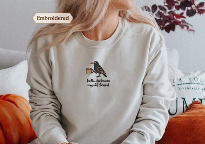 A woman wearing a white sweater with a black bird embroidered on it, sitting on a couch with a pillow behind her.