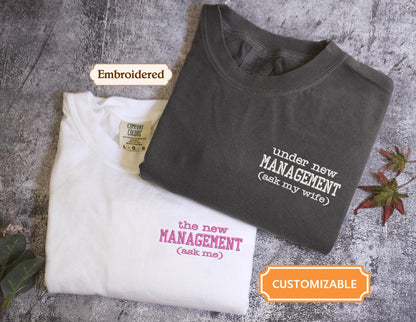 two t-shirts with humorous text printed on them, one white and one gray, placed on a textured surface.