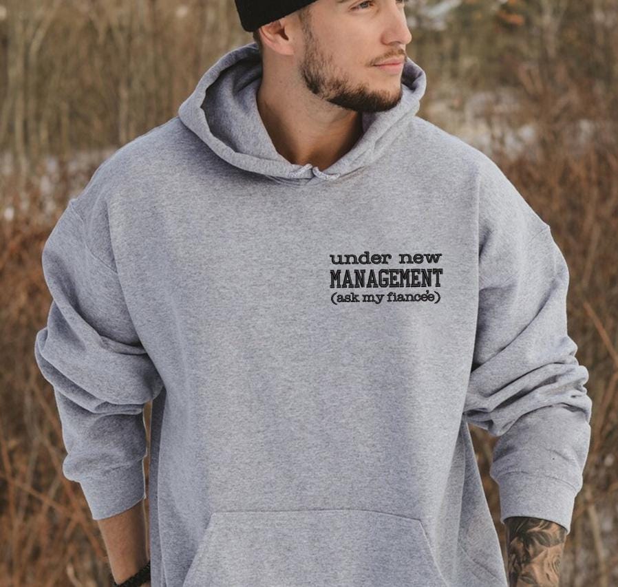 A man wearing a grey hoodie with the text &quot;under new MANAGEMENT (ask my flirts)&quot; printed on it.